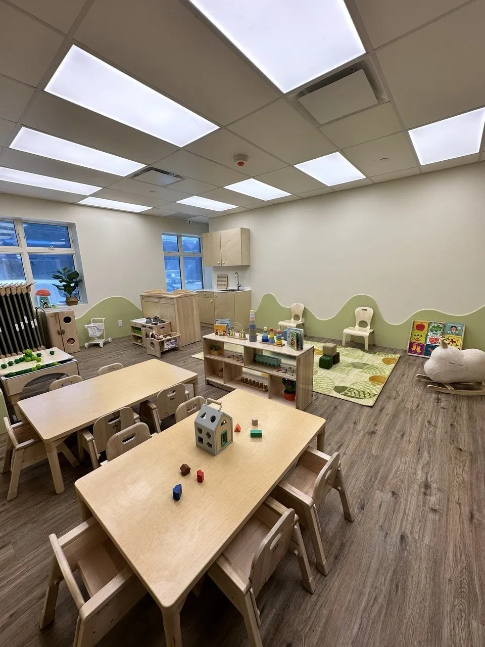 A bright classroom with small tables, shelves and a play rug